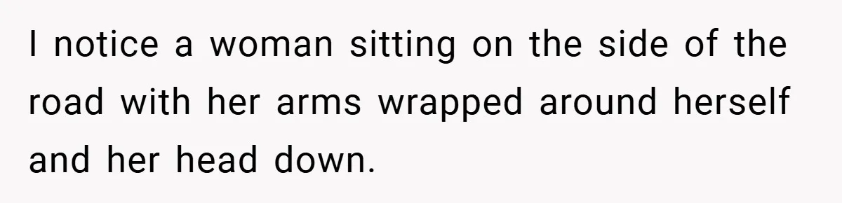 I notice a woman sitting on the side of the road with her arms wrapped around herself and her head down.