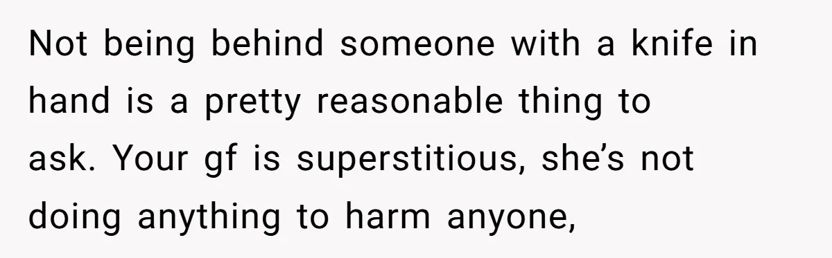 Not being behind someone with a knife in hand is a pretty reasonable thing to ask. Your gf is superstitious, she’s not doing anything to harm anyone,